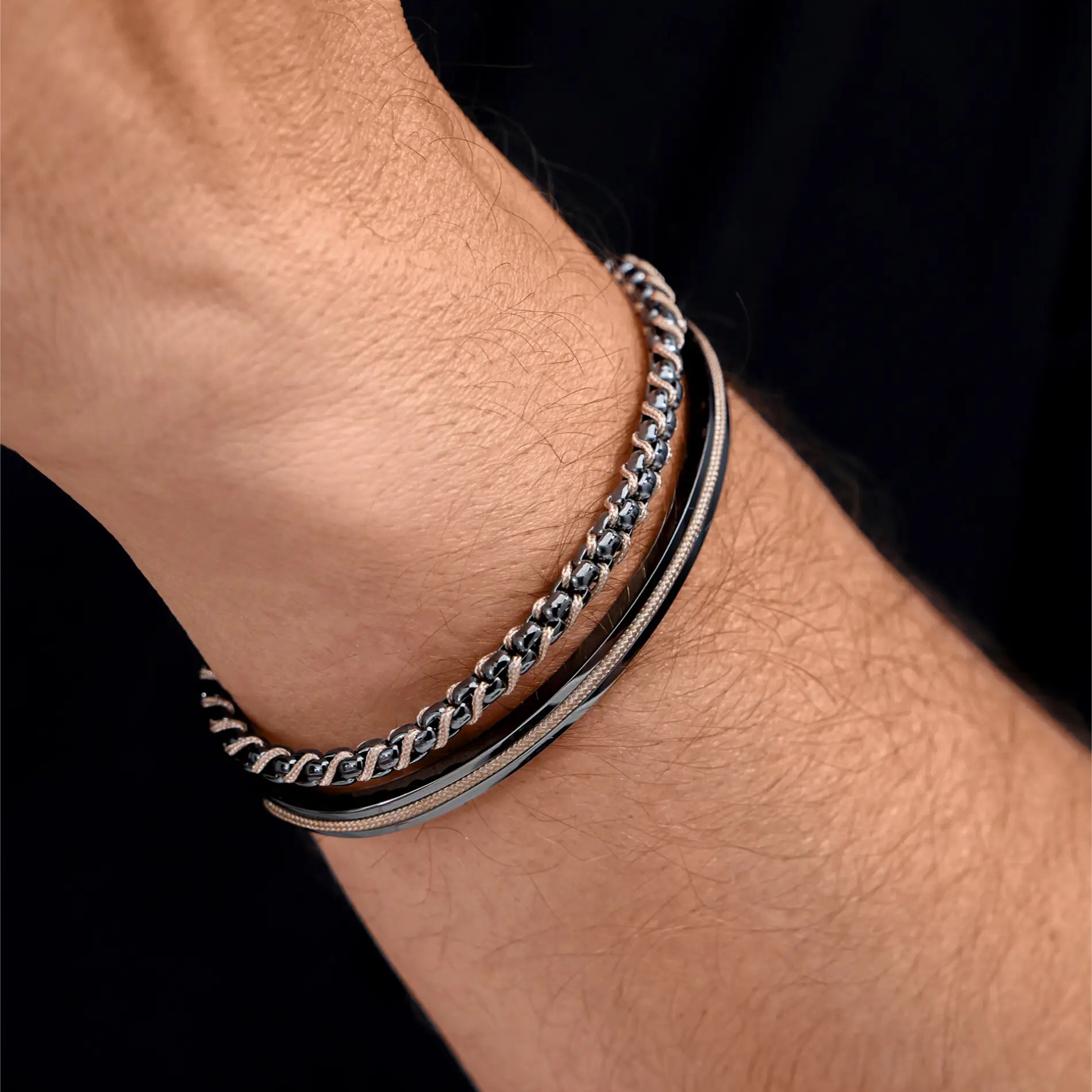 Gunmetal / Sand Rope || Man’s wrist wearing two slim intertwined bracelets featuring polished sand and black tones on dark background