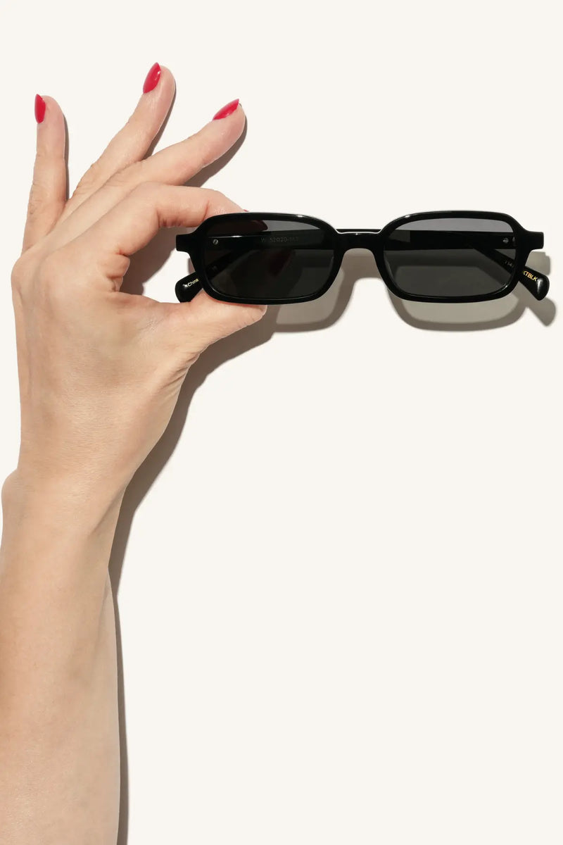 WMP Eyewear narrow rectangular sunglasses in gloss black with dark black lenses, held by a hand with red nail polish