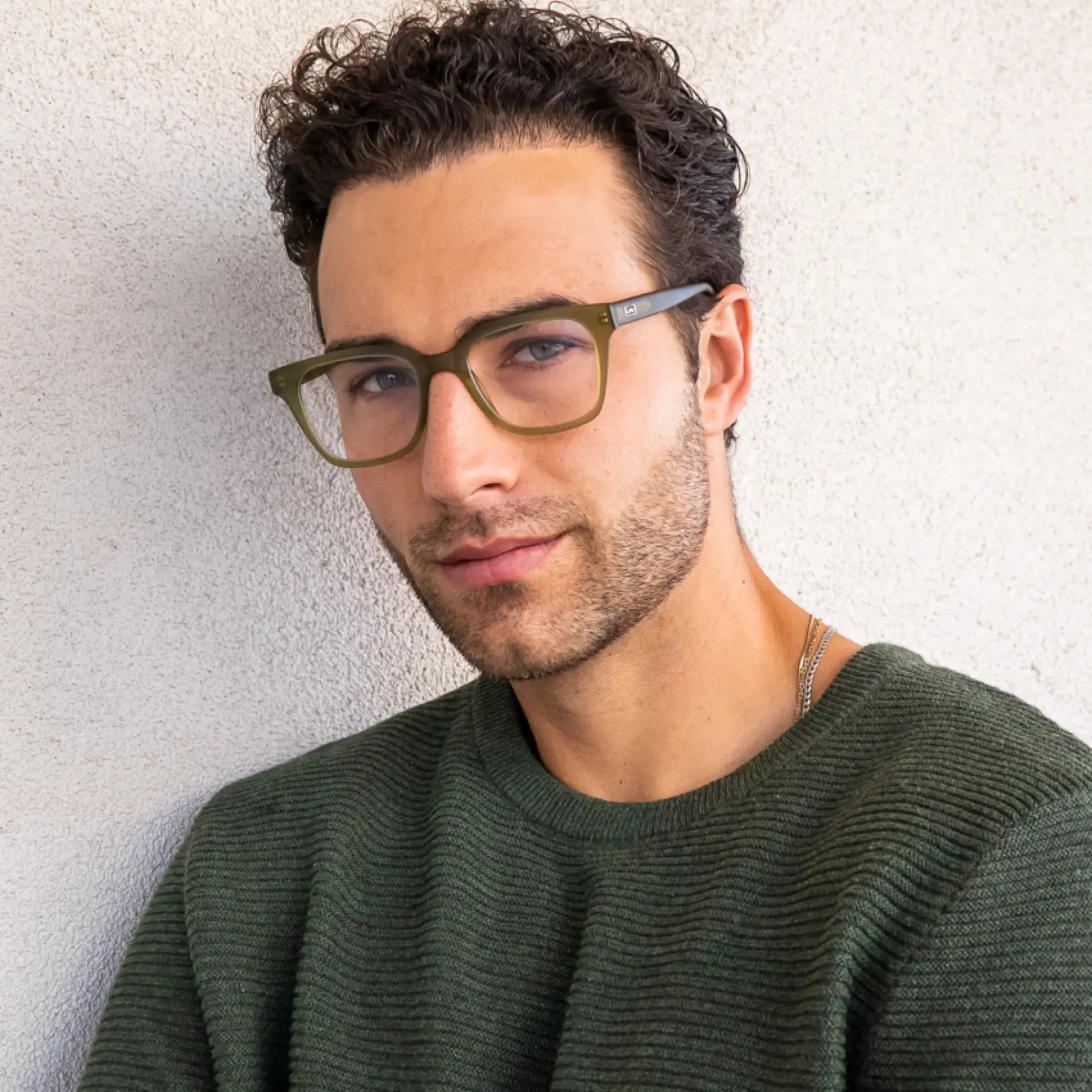 Frosted Khaki Green || Khaki green square frame blue light glasses worn by man with dark curly hair and green sweater against textured wall