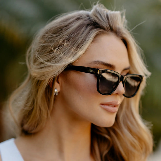 Close-up lifestyle portrait of a woman wearing black square sunglasses with dark lenses, shown in profile outdoors in soft natural light, her hair worn loose.