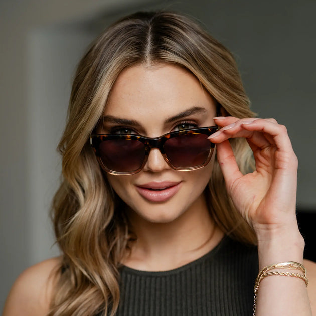 Front-facing portrait of a woman indoors wearing tortoiseshell square sunglasses with dark lenses, lifting the frames slightly with one hand and looking toward the camera.