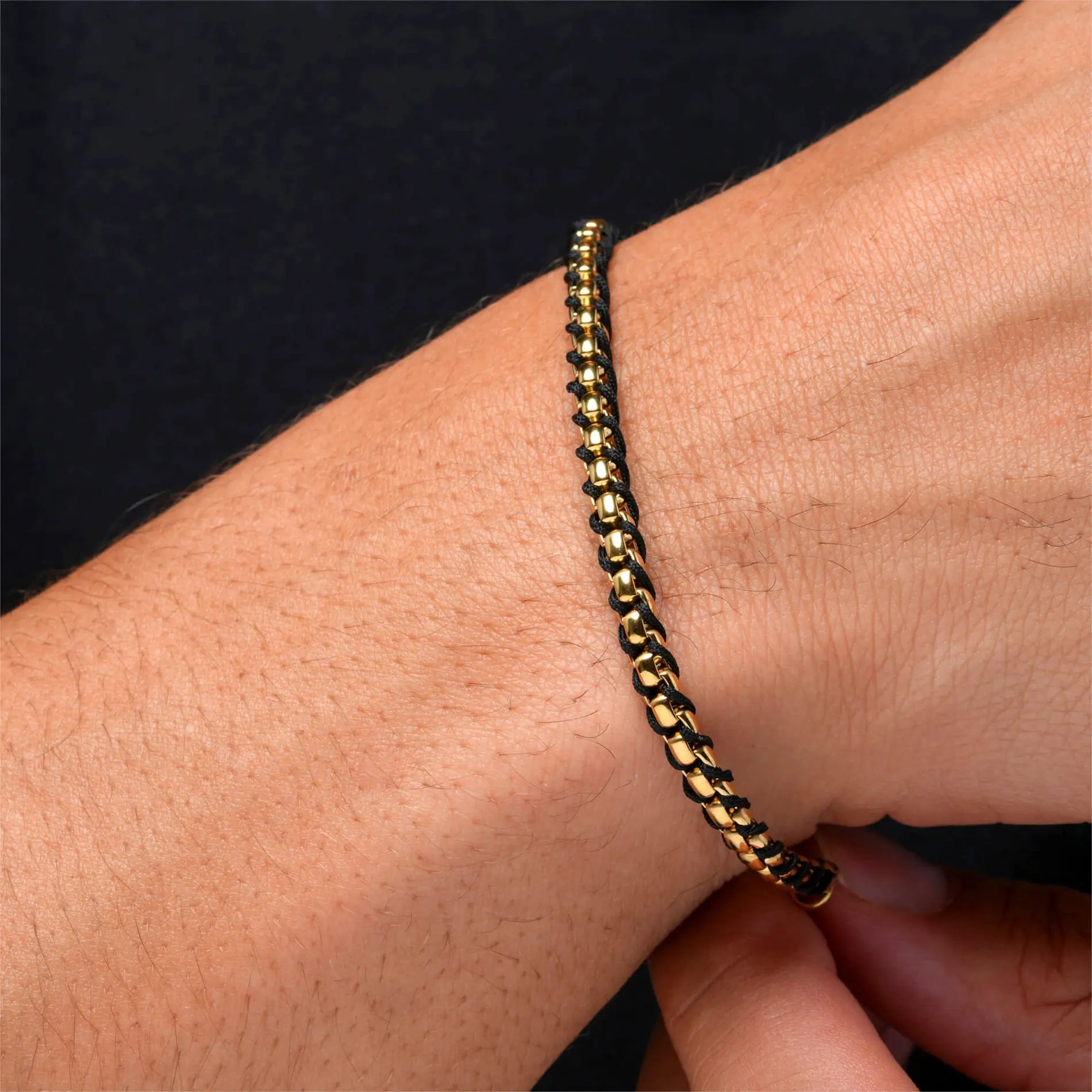 Gold / Black || Close up of wrist wearing a gold chain bracelet with black leather woven details against dark background