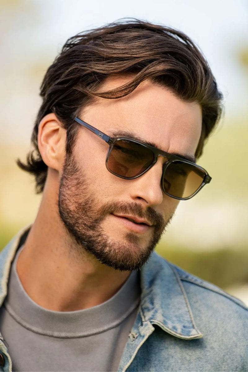 Man wearing dark rectangular sunglasses with brown lenses, looking slightly downward, dressed in a gray T-shirt and denim jacket against a softly blurred outdoor background.