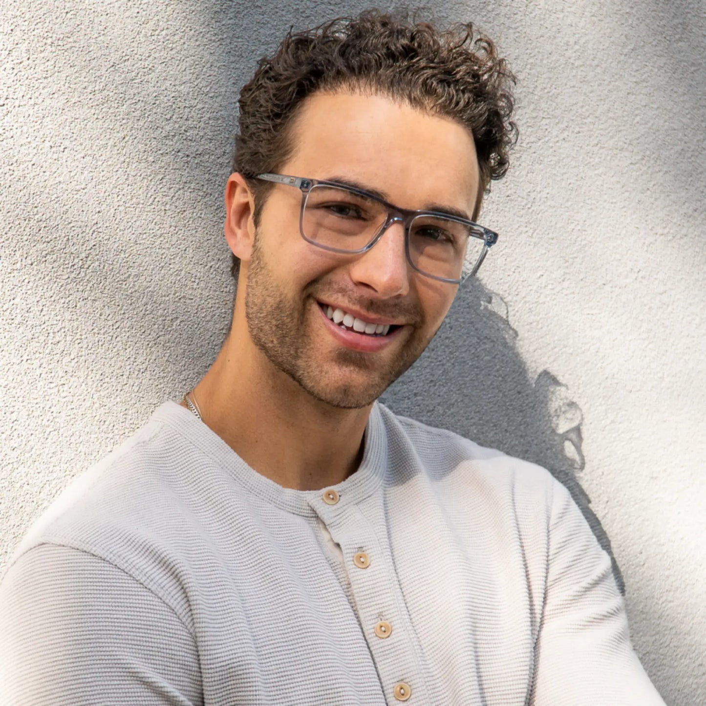 Denim Blue || Translucent gray rectangular blue light glasses worn by smiling man leaning against light textured wall