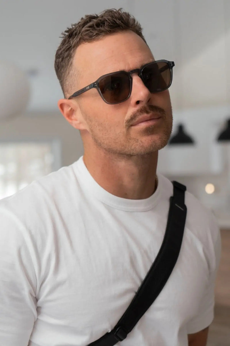 A man with short textured hair and a mustache wearing a white t-shirt and black crossbody bag strap models WMP Eyewear square sunglasses in a translucent blue frame with brown lenses, photographed looking upward in three-quarter profile with a soft indoor background.
