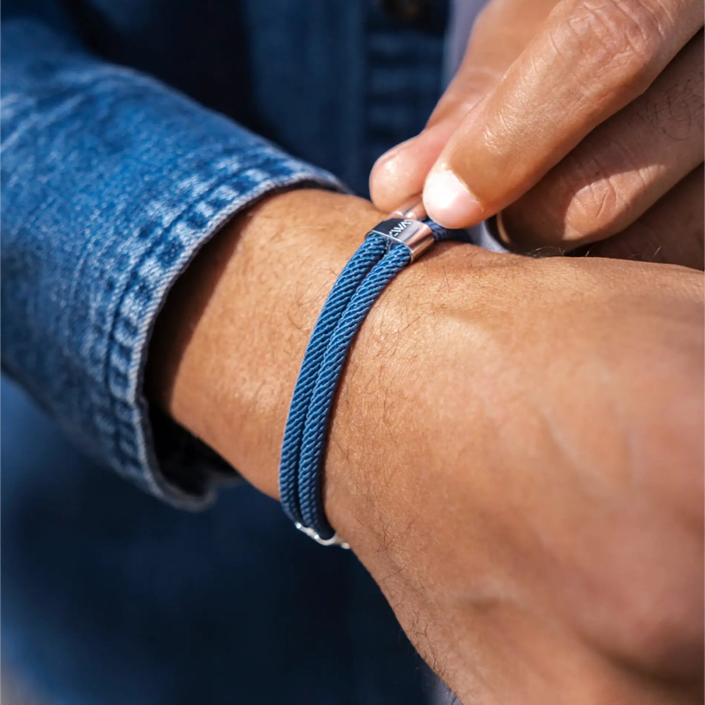 Denim Blue / Silver || Close up view of the Silver Stainless Steel Adjustable Pull Closure Blue Rope Bracelet on a man's wrist in natural lighting. 