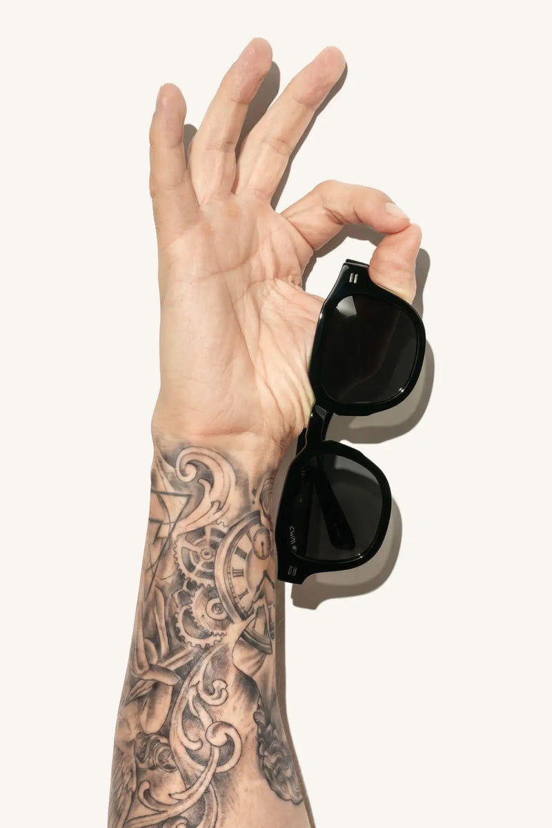 WMP Eyewear geometric sunglasses in gloss black with dark lenses, pinched between the fingers of a tattooed hand