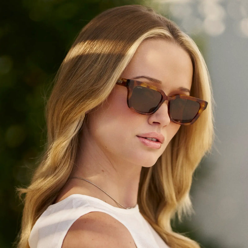 Rectangular acetate sunglasses with thick tortoiseshell frames and brown lenses worn by a woman outdoors