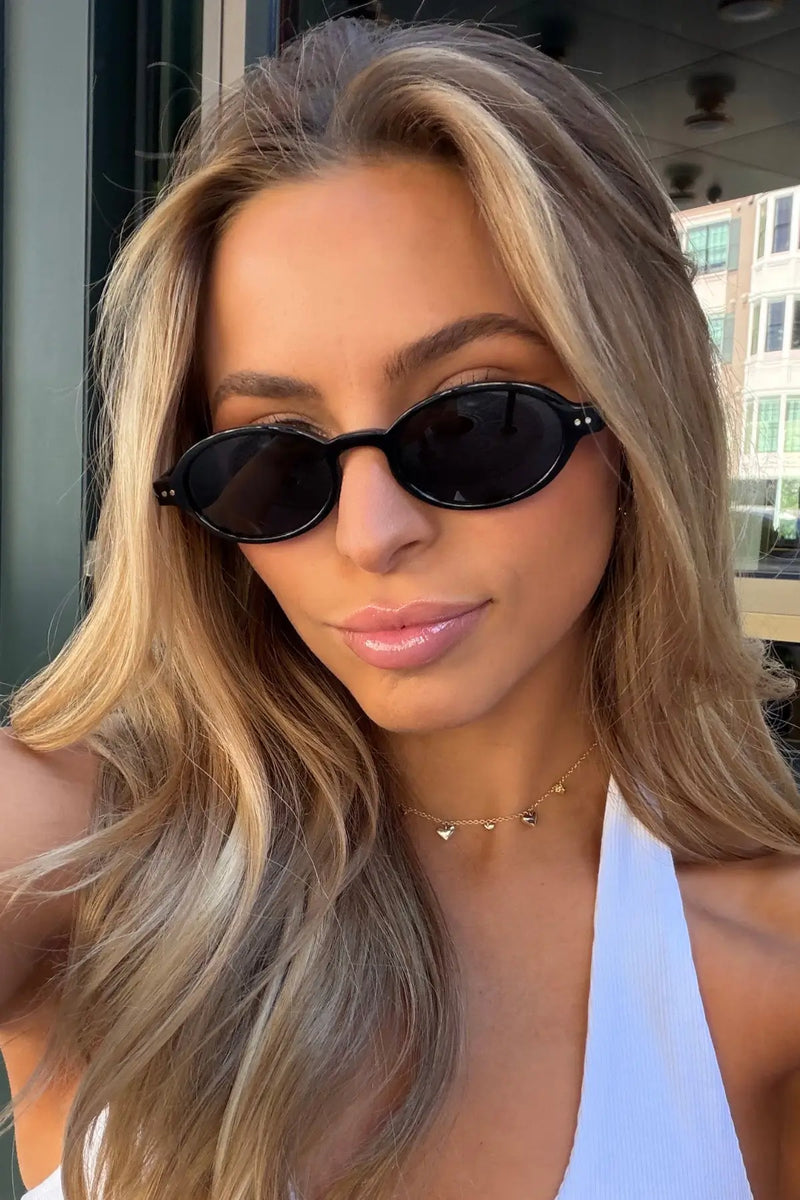 Woman wearing small black oval-frame WMP Noa sunglasses with triple black lenses, close-up selfie style.