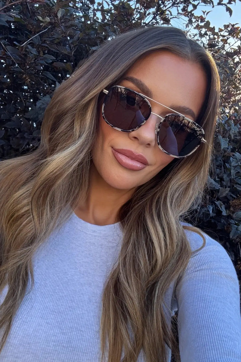 Close-up of a model wearing Gia sunglasses in Beige Tortoise outdoors with leafy trees in the background.