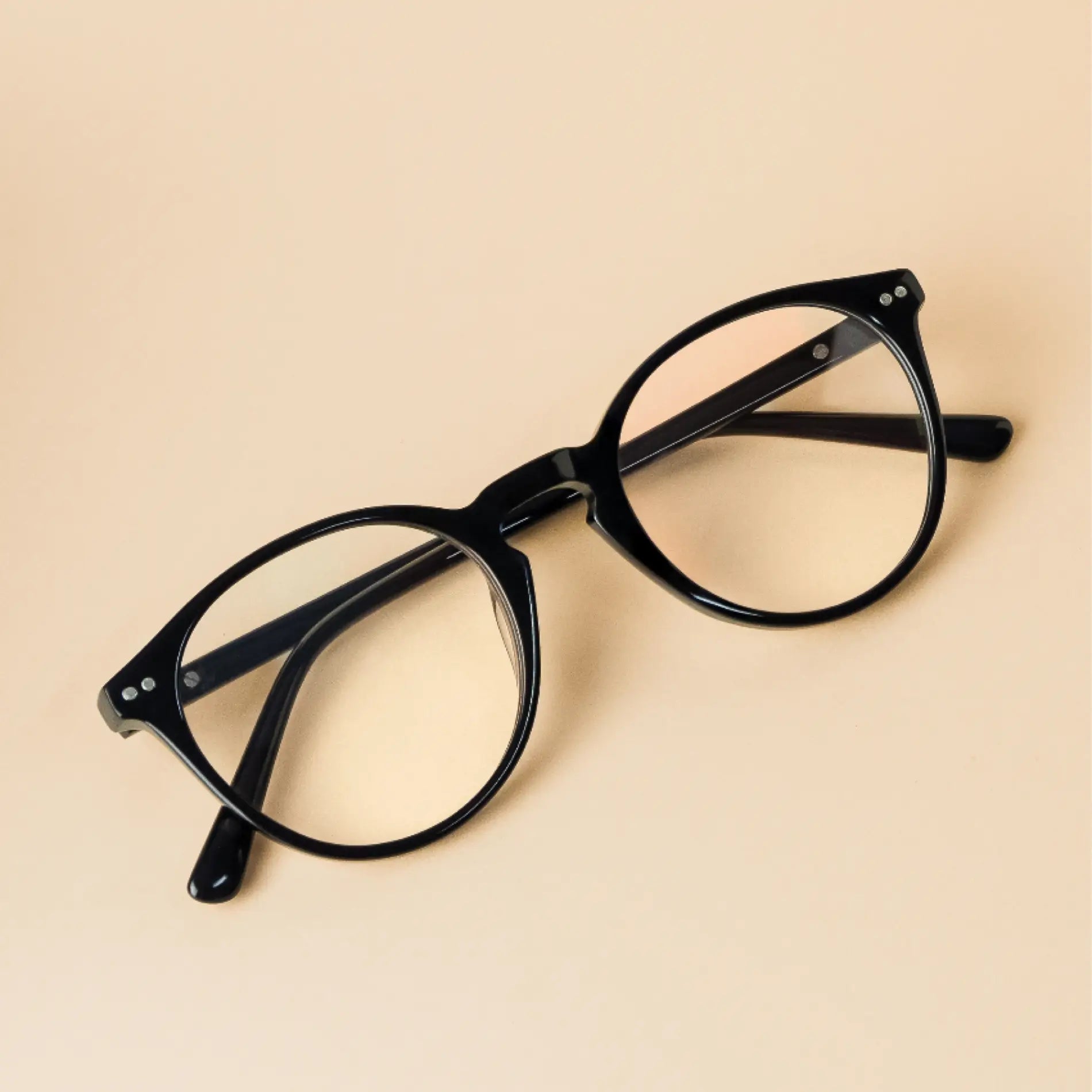 Glossy Black || Black rounded frame glasses with subtle silver accents resting on beige surface with soft lighting