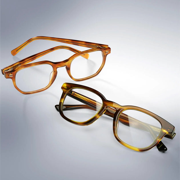 Reese eyeglasses in Honey Tortoise and James eyeglasses in Golden Brown Grain, two square acetate optical frames with clear lenses on a light grey reflective surface
