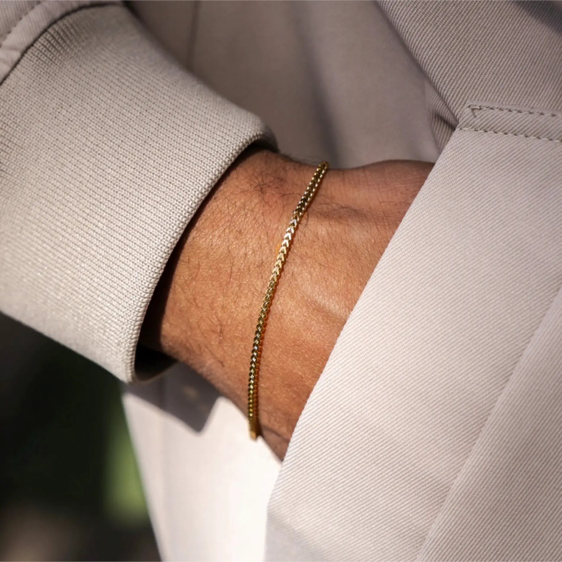 Gold || Slim gold tone bracelet with fine chain detail worn on wrist with beige suit sleeve background