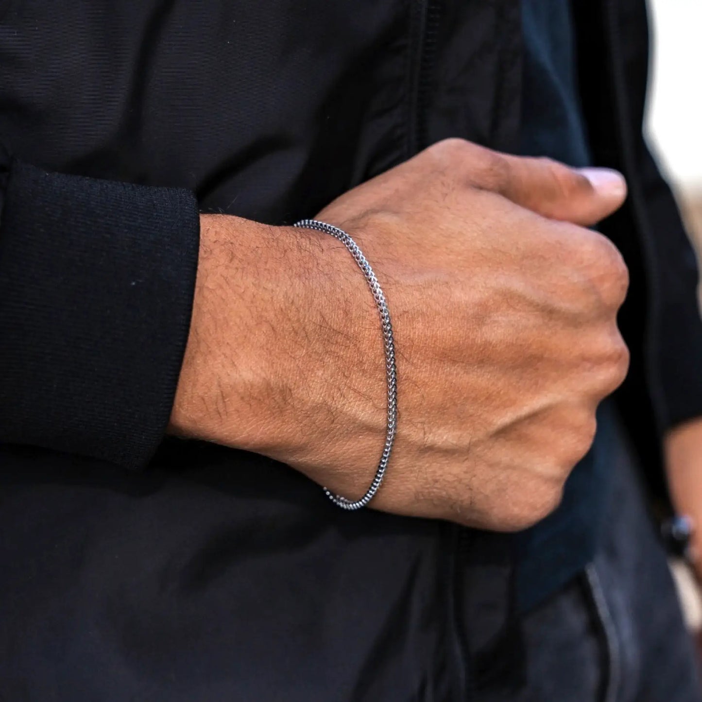 Silver || Close up of wrist wearing a slim chain bracelet with intricate weave detail over black sleeve background
