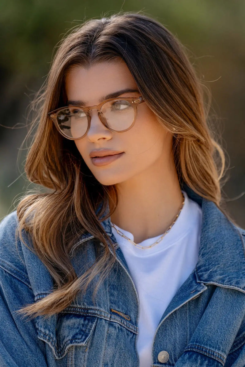Woman wearing translucent brown round eyeglasses with clear lenses, a white top, and a denim jacket, looking slightly to the side outdoors against a softly blurred green background.