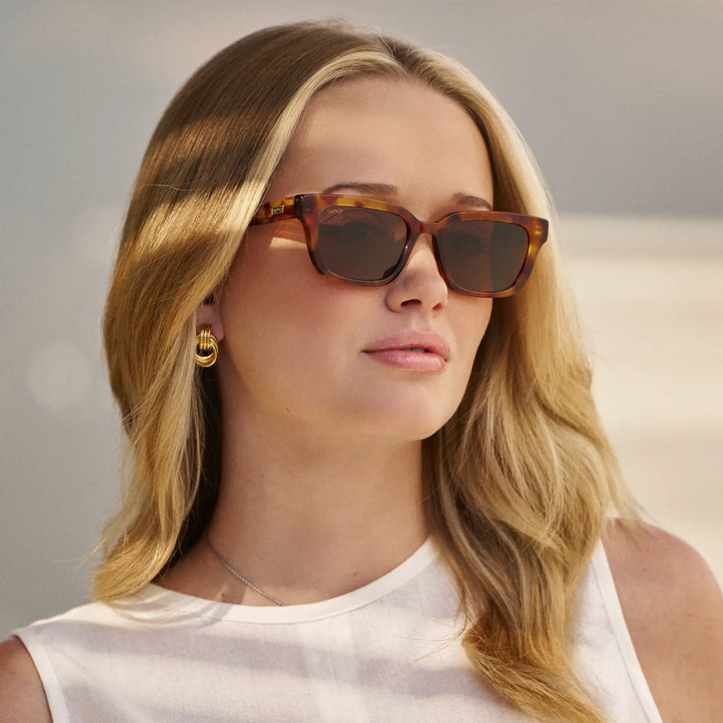 Woman wearing tortoiseshell rectangular sunglasses with dark lenses, gold hoop earrings, and a white sleeveless top, looking slightly to the side in soft natural light.