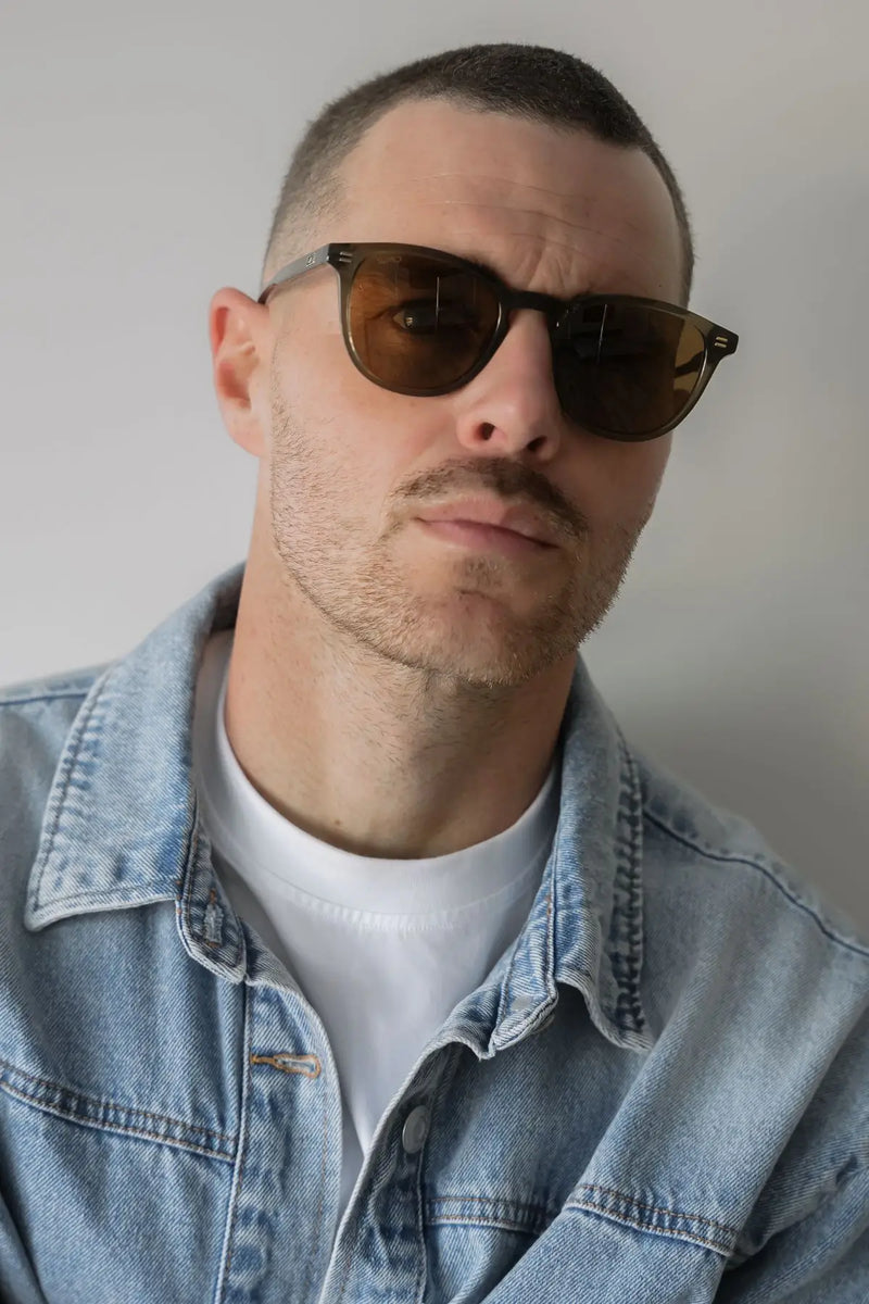 Man wearing round dark moss green WMP Walker sunglasses with brown lenses, denim jacket and white t-shirt, front view.