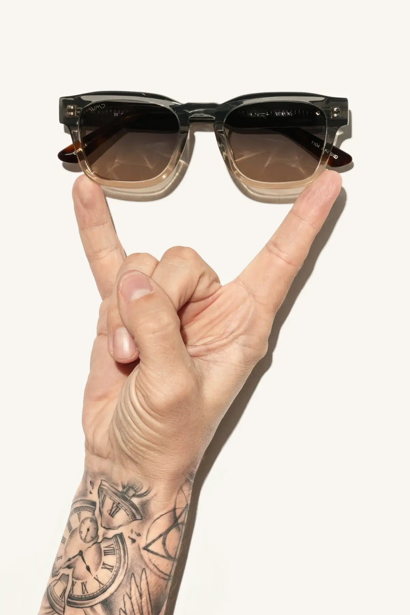 WMP Eyewear square sunglasses in two-tone graphite horizon grey and champagne with grey brown gradient lenses, balanced on two fingers of a tattooed hand