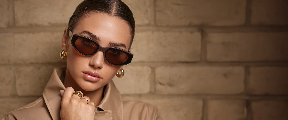 Model wearing Astra sunglasses in Mocha Brown with brown lenses, featuring a sleek, narrow cat-eye frame, styled with a beige jacket and gold jewelry against a warm-toned stone wall.