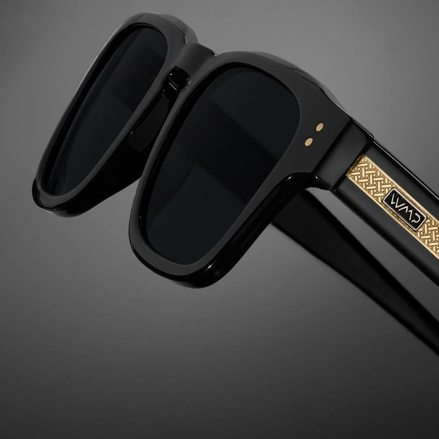 Close-up of black WMP sunglasses with gold engraved exposed temple arms against a dark gradient 