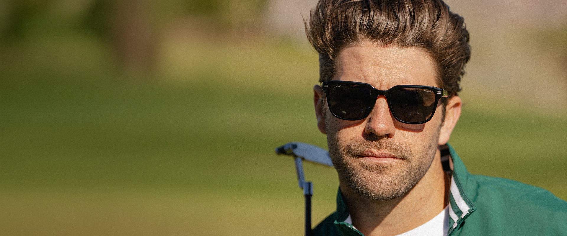 Man wearing black rectangular sunglasses on a golf course, holding a putter.
