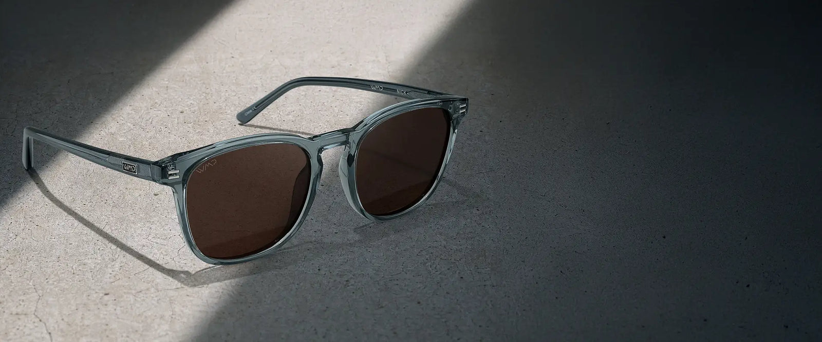 Angled product view of Nick sunglasses in Crystal Stone Blue with Brown lenses resting on a concrete surface