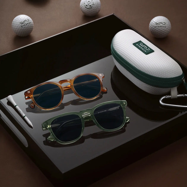 Honey tortoise and green WMP Eyewear sunglasses displayed on a black tray with a white and green golf-ball-print case, wooden tee, and WMP Eyewear golf balls