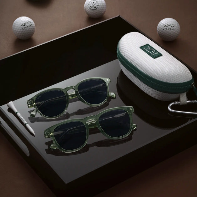 Two pairs of green WMP Eyewear sunglasses (Birdie and Wyndham styles) on a black tray with a golf-ball-print case, wooden tee, and WMP Eyewear golf balls