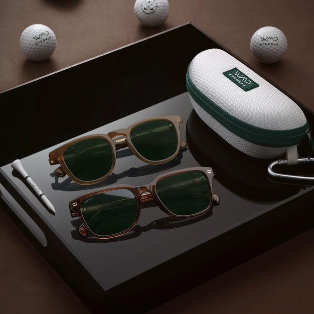 Two pairs of brown WMP Eyewear sunglasses with green lenses (Wyndham and Woods styles) on a black tray with a golf-ball-textured case, wooden tee, and WMP Eyewear golf balls