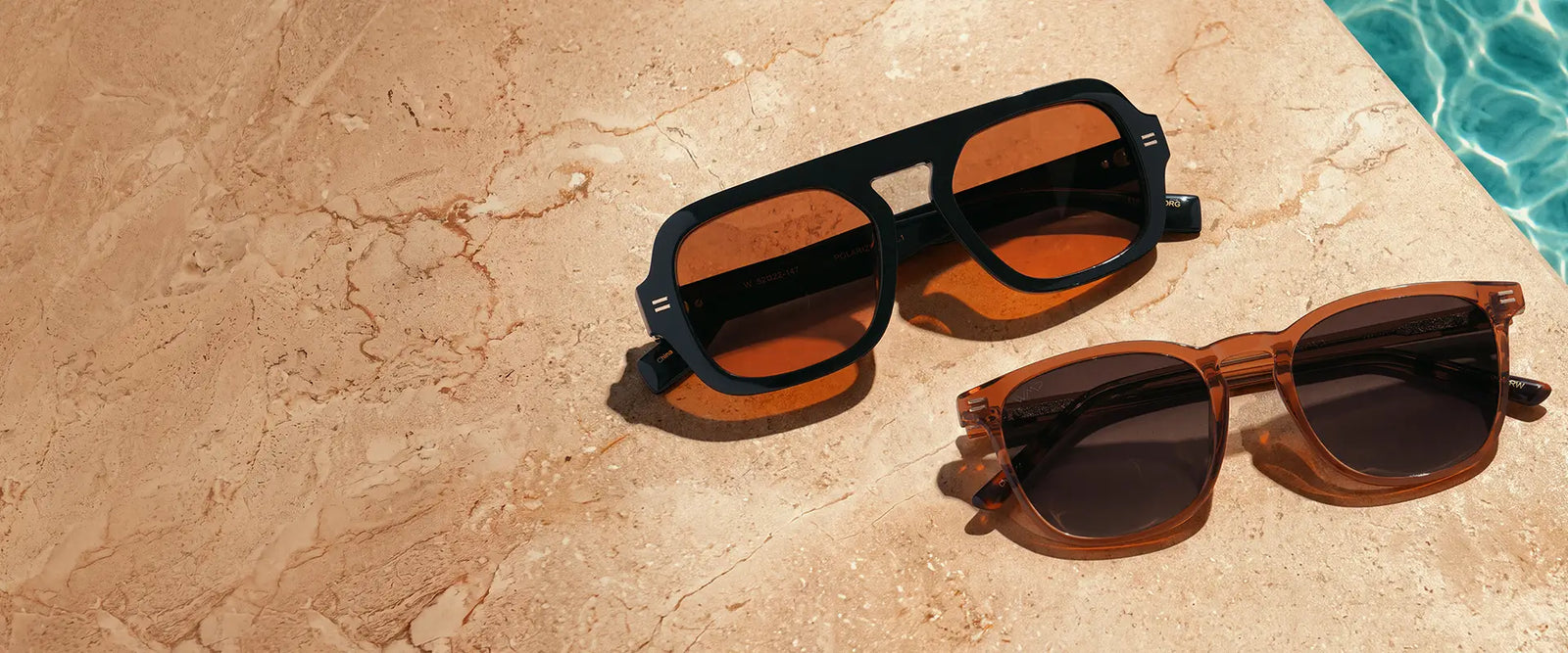 Two pairs of WMP sunglasses on marble poolside surface: brown Nick style sunglasses with brown lenses, and black Naomi aviator style sunglasses with orange tinted lenses.