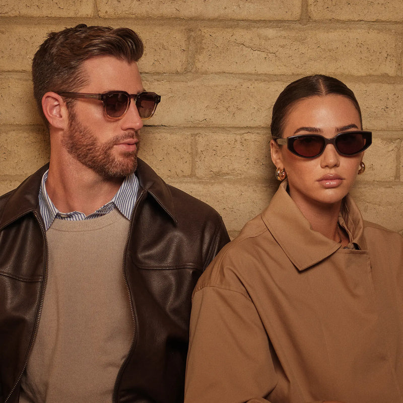 Two models wearing WMP Eyewear sunglasses in warm brown tones, styled in neutral fall layers against a tan brick wall.