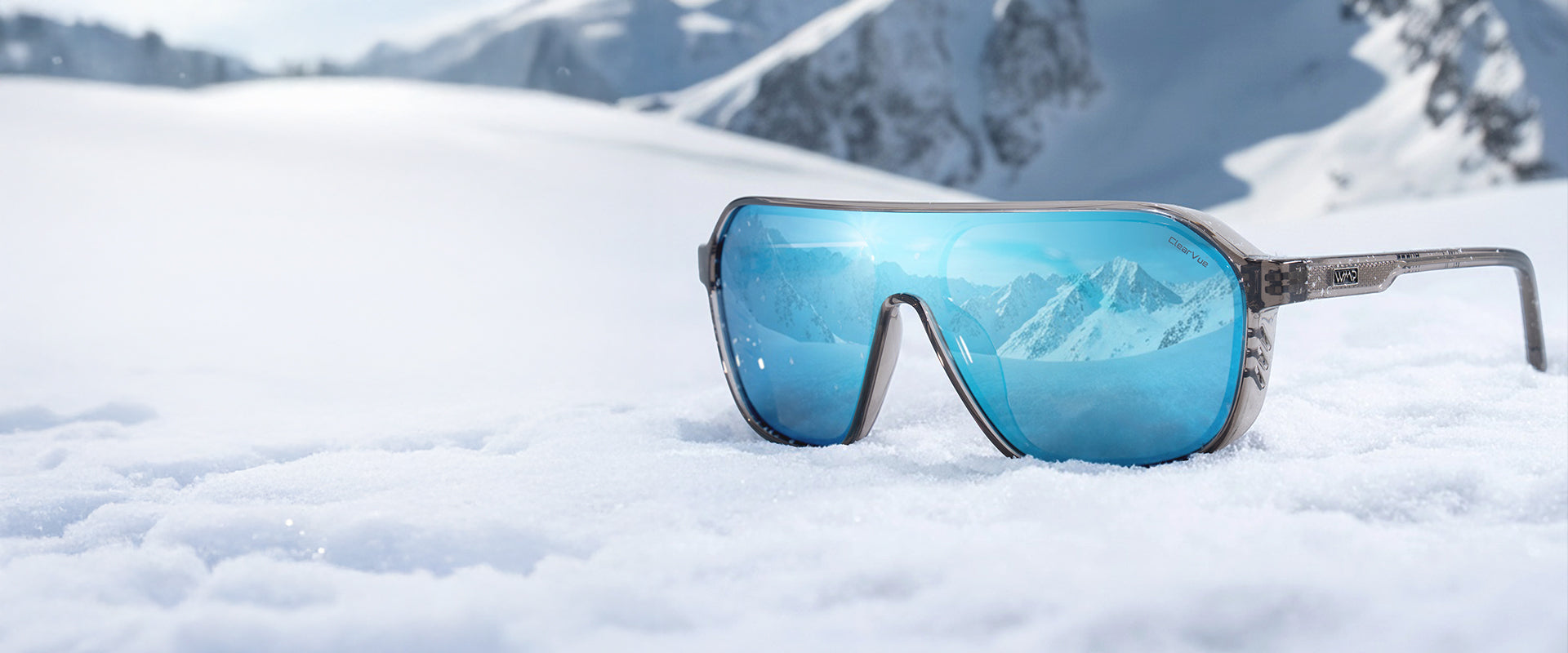 Shield-style sunglasses with blue mirrored lenses resting in fresh snow, reflecting a mountain range under a bright winter sky.