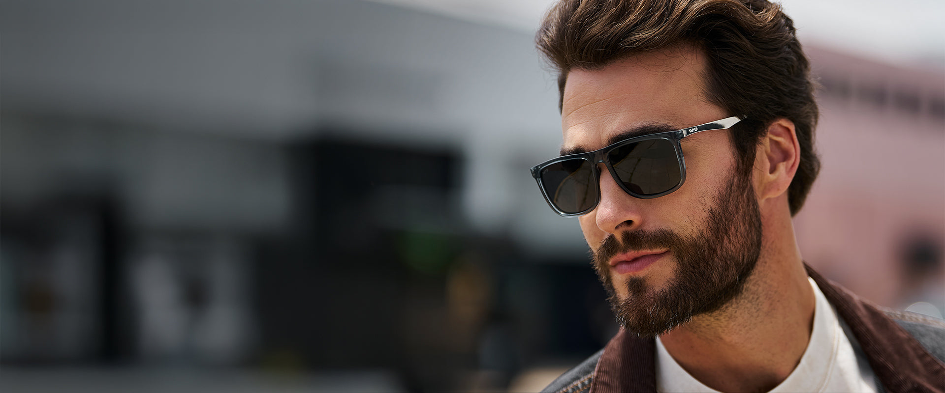 A man with short brown hair and a full beard wears rectangular blue sunglasses with slim silver-accented temples. He is outdoors, looking off to the side, with a softly blurred urban background behind him.